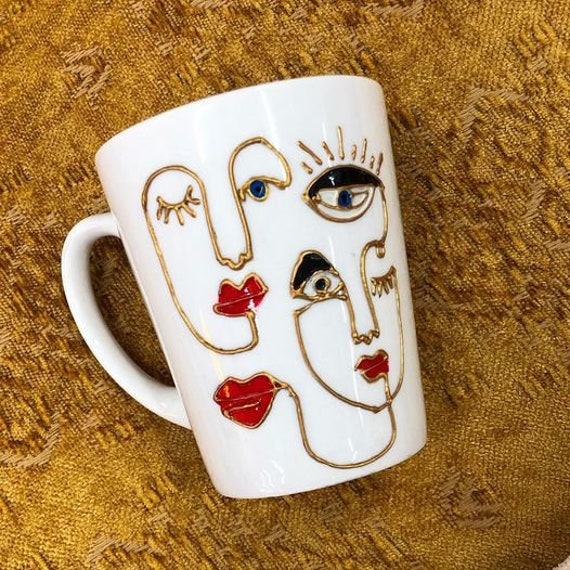 Weiße Tasse bemalt mit abstrakten Gesichtern in goldenen Linien und roten Lippen