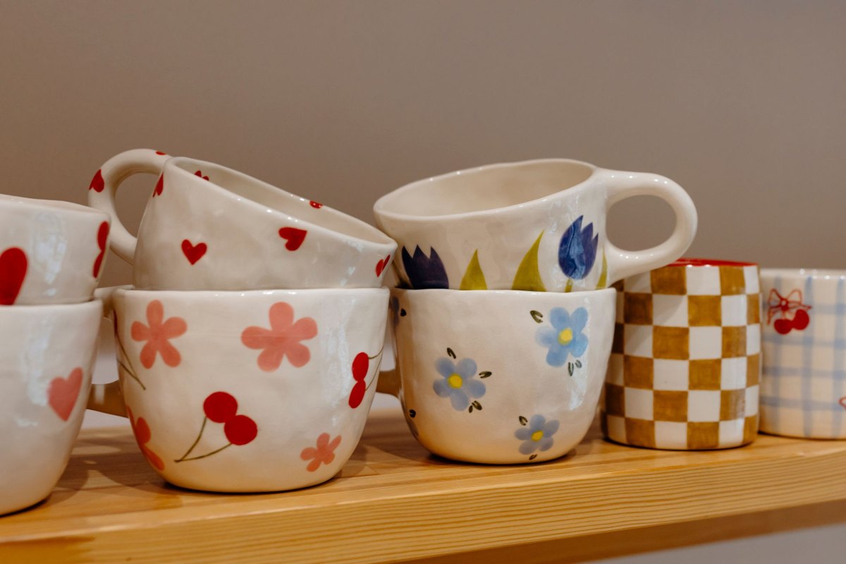 Handbemalte Keramikbecher mit roten Kirschen, rosa Blüten und Karomustern auf einem Holzregal
