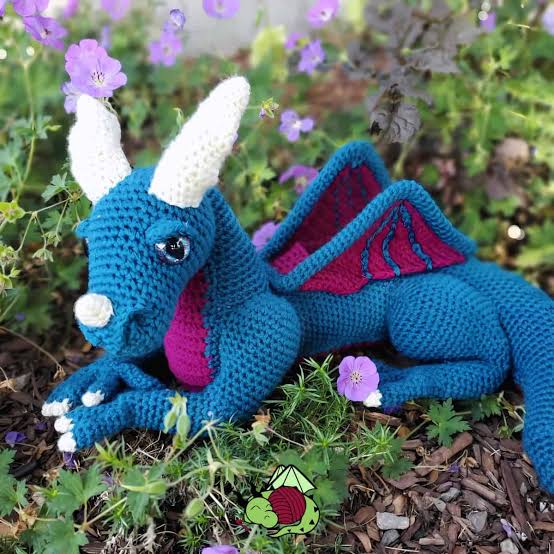 Leuchtend blauer Häkeldrache mit weißen Hörnern und pinken Flügelinnenseiten zwischen Blumen