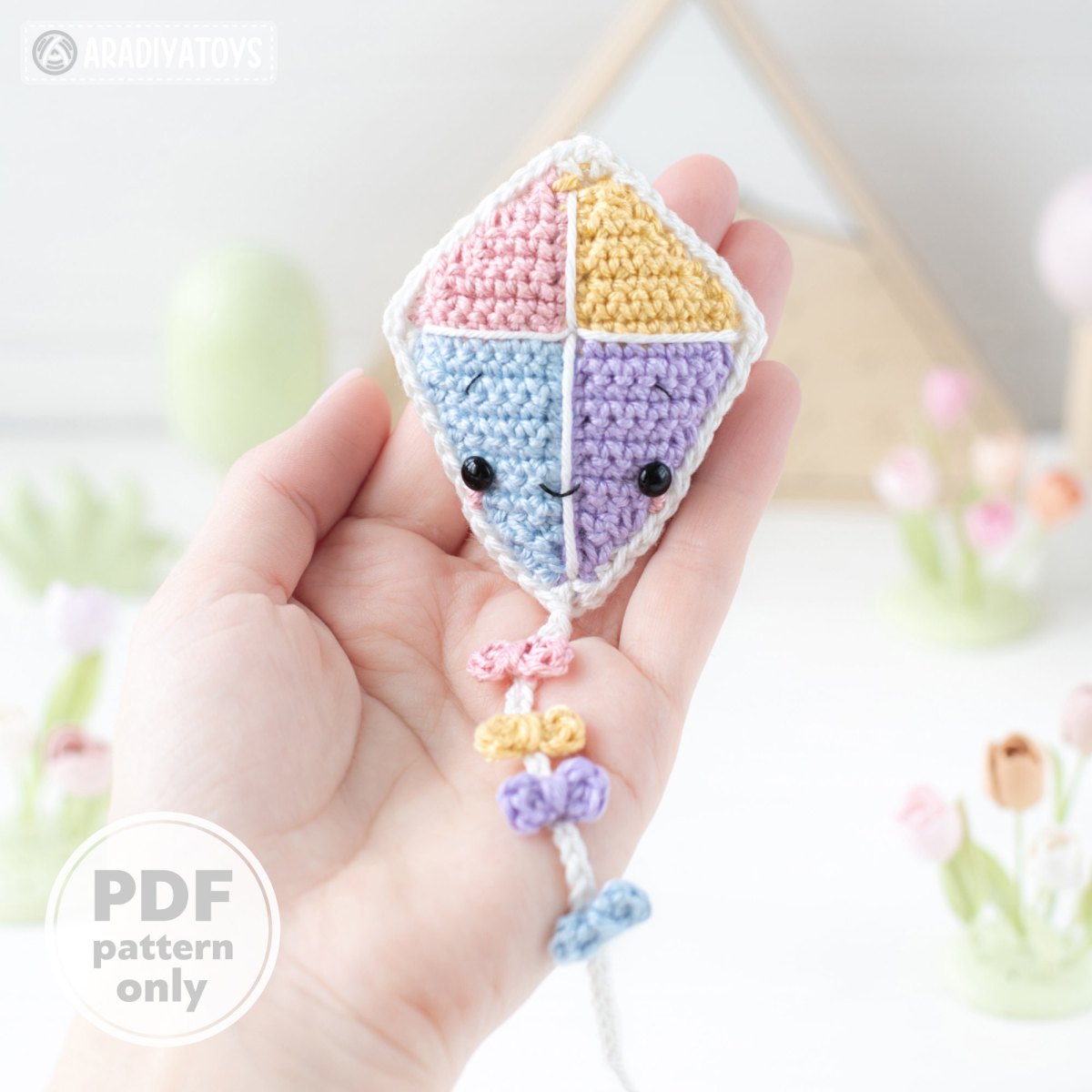 Hand hält einen kleinen pastellfarbenen gehäkelten Flugdrachen mit Kawaii-Augen und kleinen Schleifen