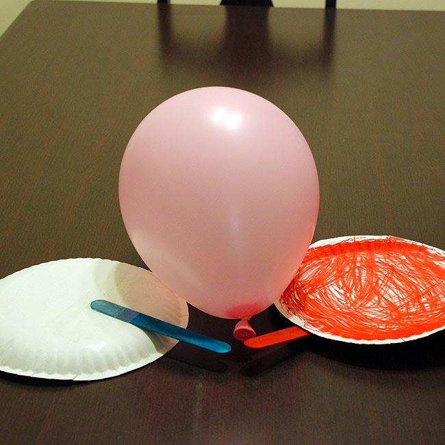 Rosa Ballon am Strohhalm zwischen zwei Papptellern, einer mit roten Kritzeln, dazu ein blauer Kunststoffstab