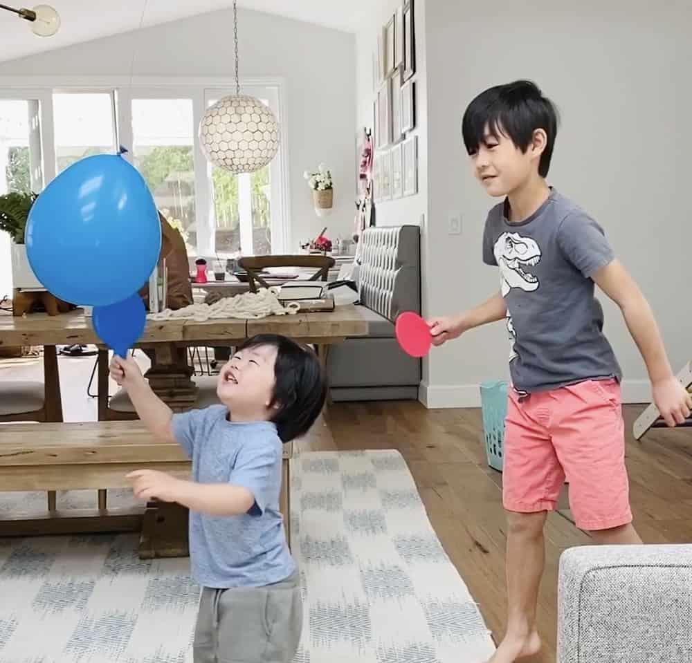 Zwei Jungen im Wohnzimmer: Der kleinere hält einen blauen Ballon oben, der größere hat einen runden roten Schläger