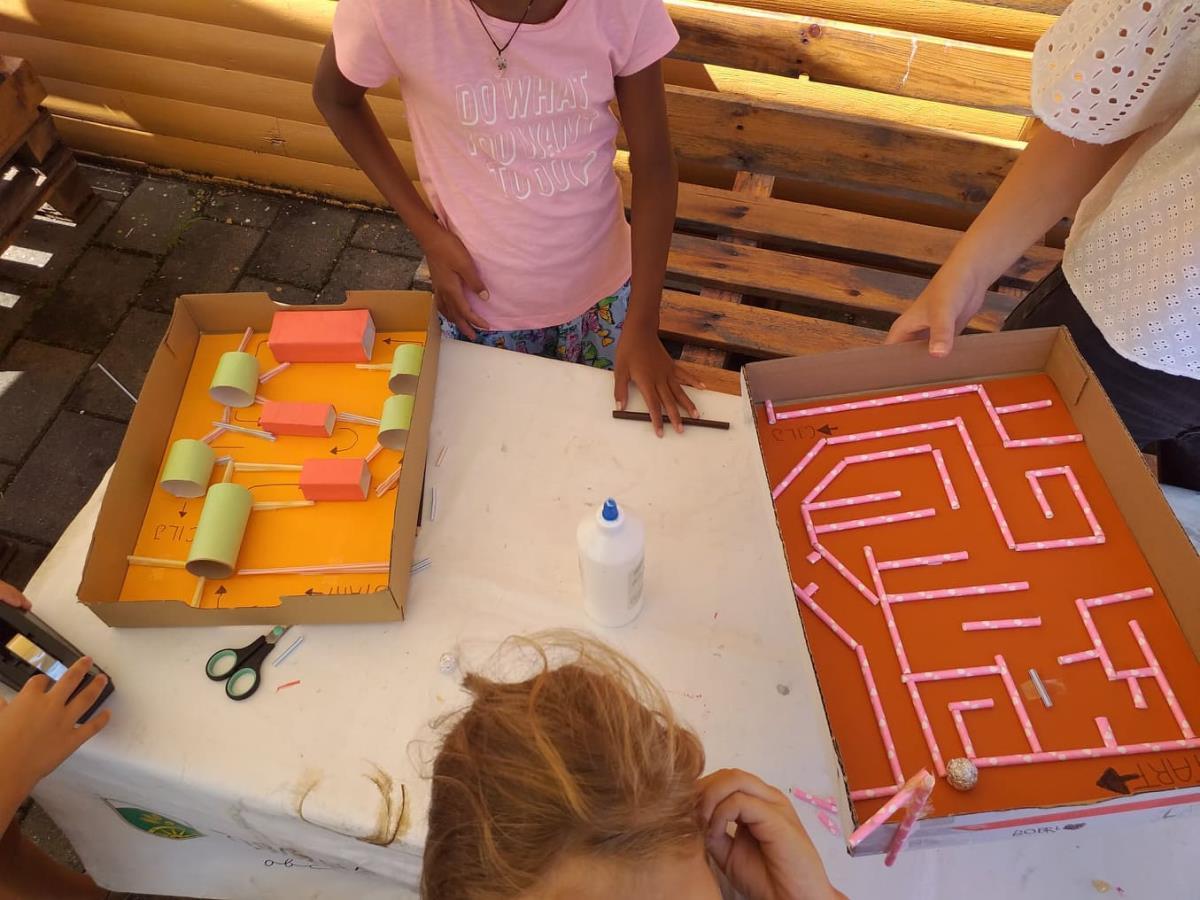 Zwei Kartons mit orange bemalter Basis: links Röhren und Klötze als Hindernisse, rechts ein Labyrinth aus rosa Strohhalmen oder Streifen, Kinder arbeiten am Tisch mit Kleber und Schere
