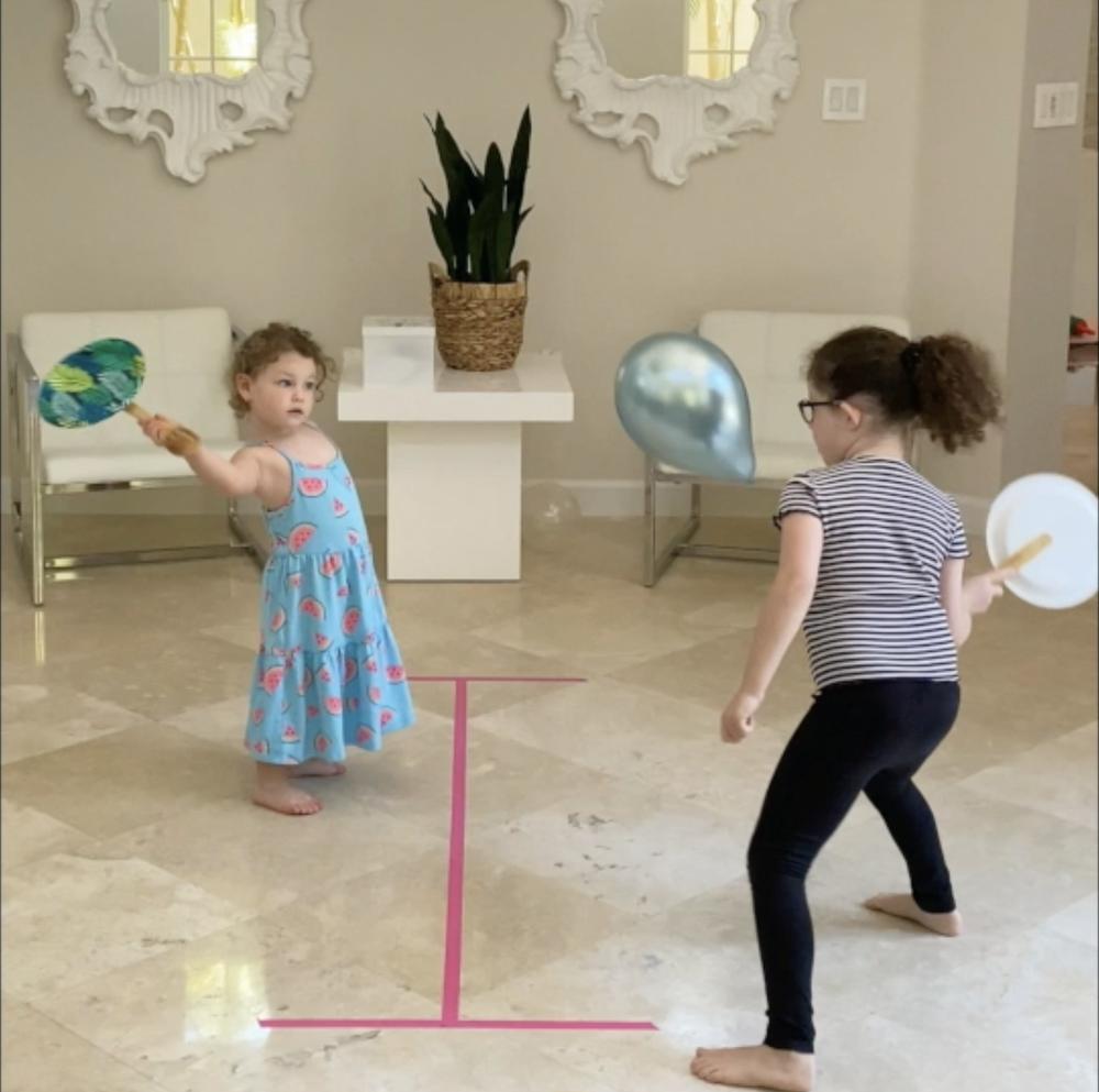 Zwei Mädchen spielen mit Papp-Schlägern und einem silbernen Ballon, auf dem Boden markiert eine rosa Klebebandlinie das Spielfeld