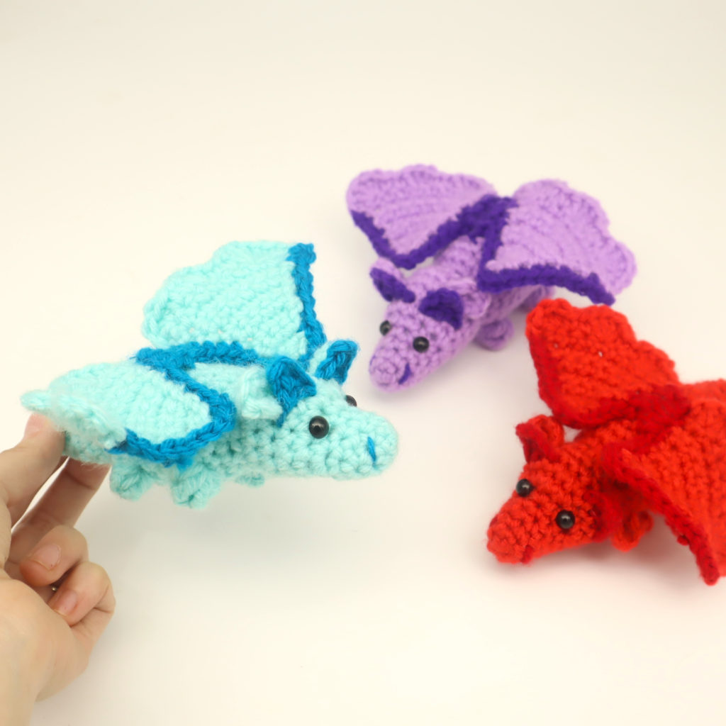 Drei kleine gehäkelte Mini-Drachen in Hellblau, Lila und Rot neben einer Hand